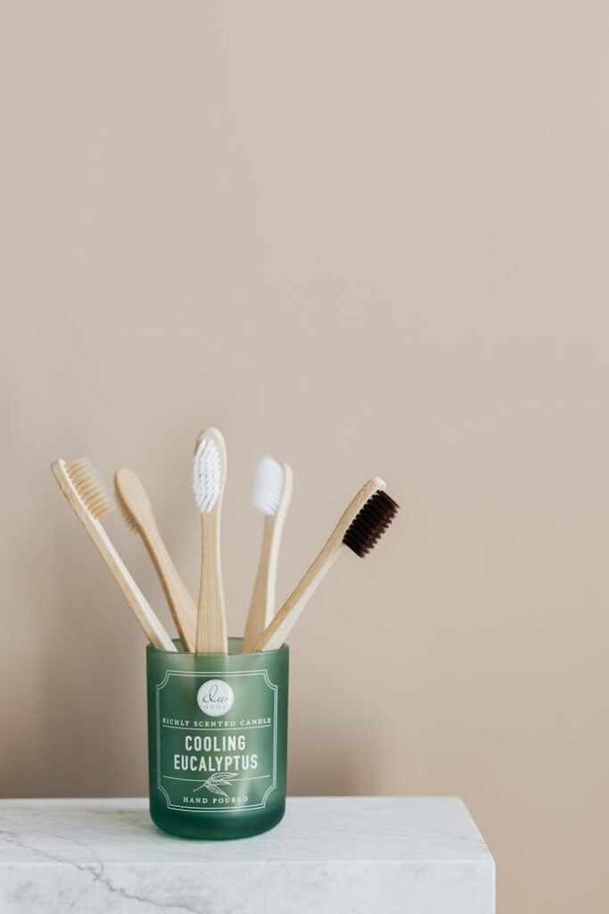 Set of various environmentally friendly toothbrushes placed in translucent green lass holder on white ceramic table against beige wall